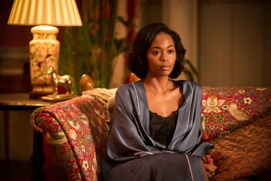 Woman in a blue robe sits on a red floral sofa in a warmly lit room, looking thoughtful.