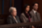 Three people sit solemnly in a courtroom setting. They are wearing suits, with dark and neutral tones. The mood appears serious.