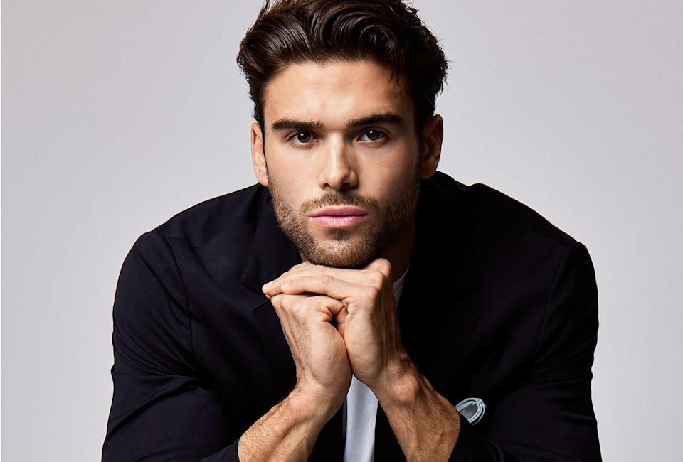 Man with beard in a black suit rests chin on hands, looking intently at the camera. Light gray background, calm expression.