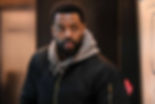 Bearded man in a black jacket and gray hoodie indoors, looking alert and concerned in a softly lit hallway.