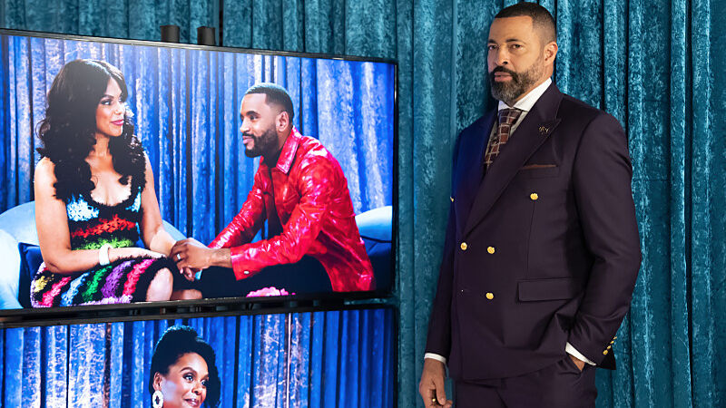 Man in a dark suit stands beside TV screens showing a couple, against blue curtains, with a serious expression.