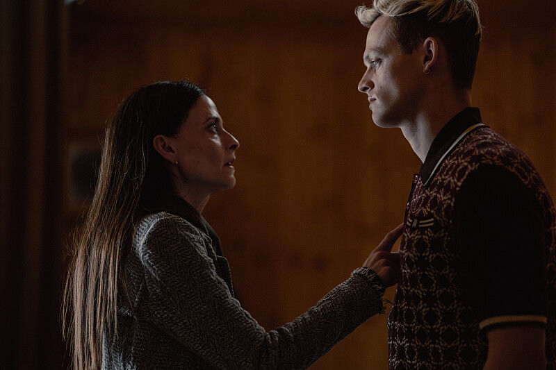 A woman intensely grabs a man's shirt in a dimly lit room with wooden walls. Tension is evident as they face each other closely.