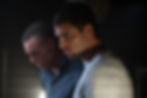 Two men in a dimly lit room, one in a gray shirt looks serious. Background shadowy and blurred, minimal colors, tense mood.