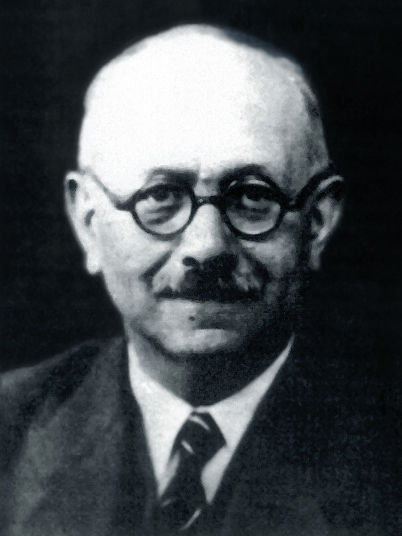 Portrait noir et blanc d’un homme chauve à lunettes rondes, en costume sombre, sur fond noir, regard neutre.