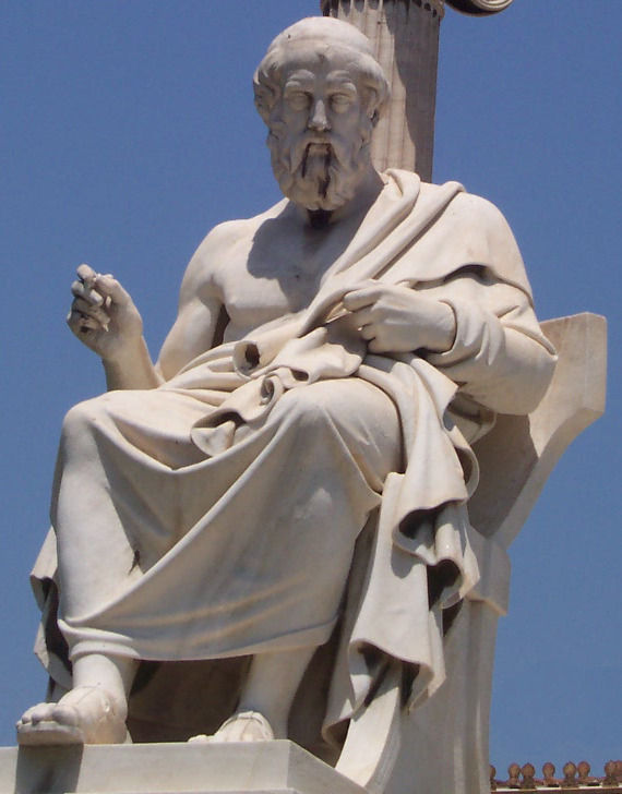 Statue en pierre blanche d’un homme barbu assis, drapé, sur fond de ciel bleu.