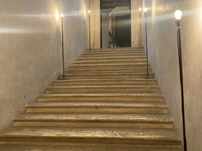 Escalier en pierre dans un couloir étroit, éclairé par des lampes, menant à une porte; ambiance calme et ancienne.