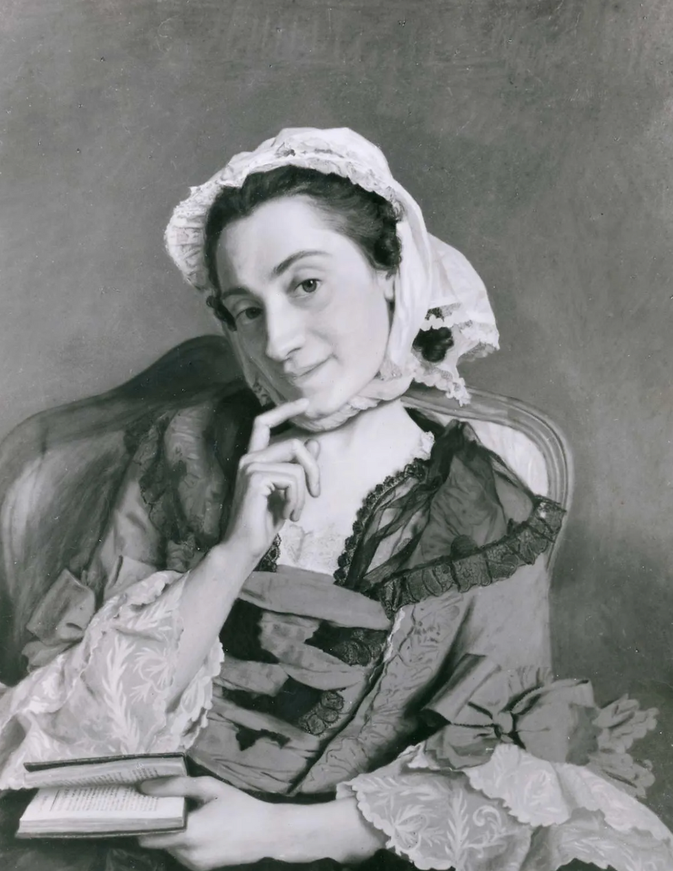 Portrait en noir et blanc d’une jeune femme en bonnet, songeuse, doigt au menton, tenant un livre.
