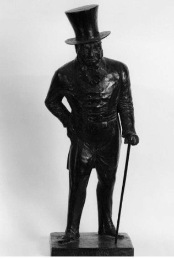 Statue noire d’un homme en haut-de-forme, appuyé sur une canne, debout sur un socle devant un fond blanc.