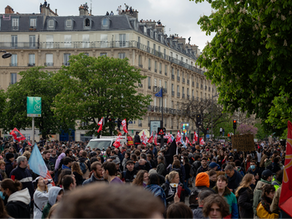 Foule manifestant dans une rue parisienne avec drapeaux rouges et pancartes. En arrière-plan, immeubles haussmanniens et arbres verts.