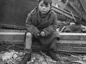 Un enfant assis sur des ruines, portant un manteau et des chaussures usées. Regard triste, fond de débris en bois et gravats. Noir et blanc.