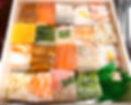 A wooden box displays assorted square sushi pieces topped with various fish and vegetables. Vibrant colors and a plastic leaf garnish complete the scene.
