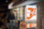People outside a convenience store at night, with a glowing "Seven & i Holdings" sign. A no-cars sign is visible. Moody ambiance.