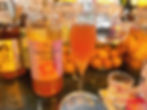 A tall glass of orange Cinnamon Umeshu next to a labeled shochu bottle on a bar counter with jars of fruit in the background. Vibrant and inviting.