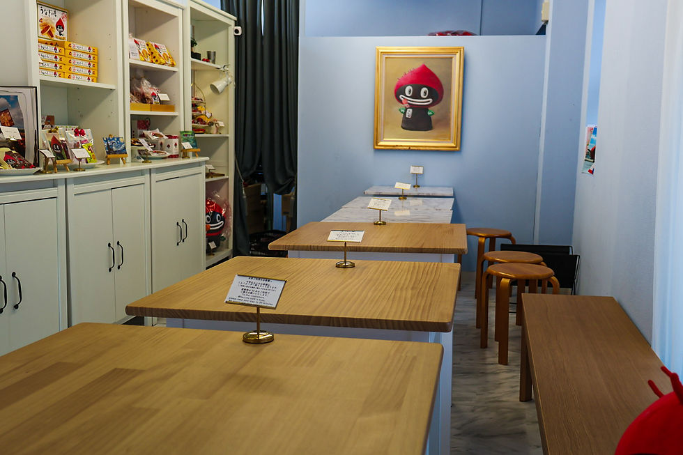 Interior of the Unagiimo cafe with wooden tables, white shelves displaying snacks, a whimsical framed painting on blue walls, and informational signs on tables.