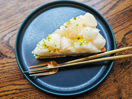 A slice of pear and pistachio tart on a blue plate with a fork and spoon, placed on a wooden table.