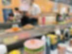 Sushi conveyor belt with colorful plates and food items; people dining in the background. Lively atmosphere.