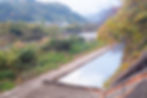 Path with railing by a canal, surrounded by green foliage and autumn leaves. Hills and houses are visible in the misty background.