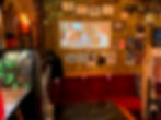Cozy interior of the bar with red velvet seating, a movie projected on a wall with subtitles, surrounded by framed pictures and posters. Plants on the side.