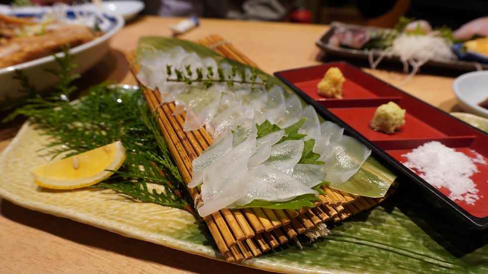 Sliced sashimi arranged on a bamboo mat with greenery, lemon wedge, and divided dish of wasabi, ginger, and salt on a textured plate, in a dining setting.