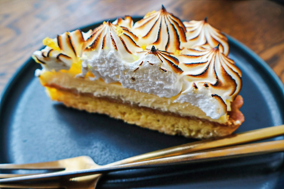 A slice of lemon meringue tart with golden-browned peaks on a black plate. Wooden table background and two gold forks beside it.