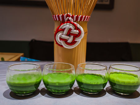 Four glass cups with green tea (matcha) on a table. Behind is a decorative bamboo arrangement with red and white cord arranged in an intricate mizuhiki design. Calm indoor setting.