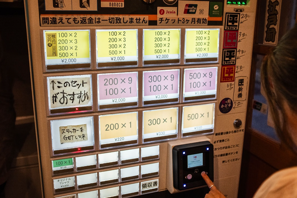 A person uses a vending machine with buttons displaying prices and Japanese text. Brightly lit display in a dimly lit indoor setting.