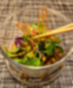 A glass bowl with fresh greens, edible butterfly made with burdock root held by chopsticks. Placed on a woven mat, evoking a sophisticated dining scene.