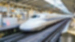 White bullet train at a busy station platform, blue stripe along its side. Passengers nearby, digital sign shows number "009". Bright, clean setting.