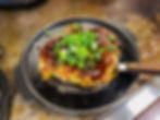 Okonomiyaki topped with chopped green onions and sauce on a hot iron skillet. Wooden spatula on the side. Warm, rustic setting.