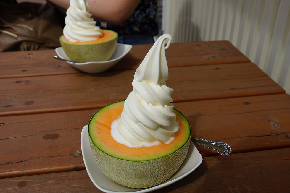 Soft-serve ice cream swirls atop halved Japanese melons on white plates and wooden table; visible hand and patterned shirt in background.