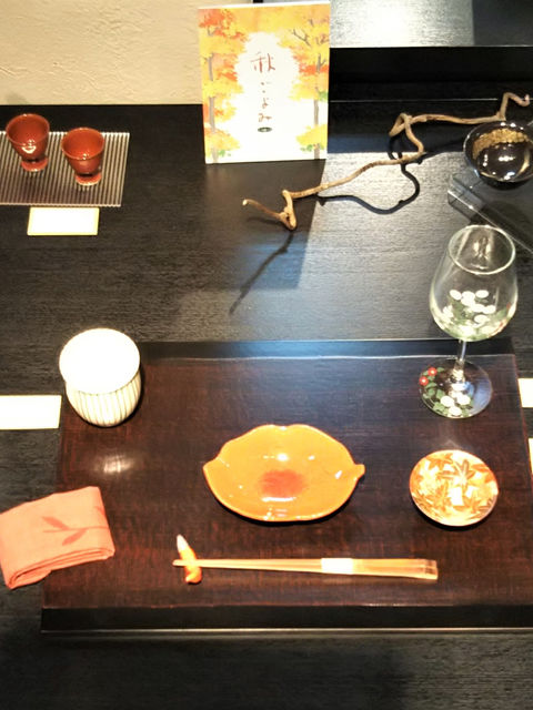 秋の展示。黒い台に朱色の丸皿や赤い徳利、ぐい呑、紅葉を並べた様子