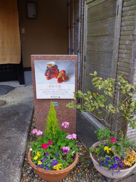 「酒器百撰」のポスターを掲げた玄関先の看板と、パンジーや黄色い花を植えた鉢