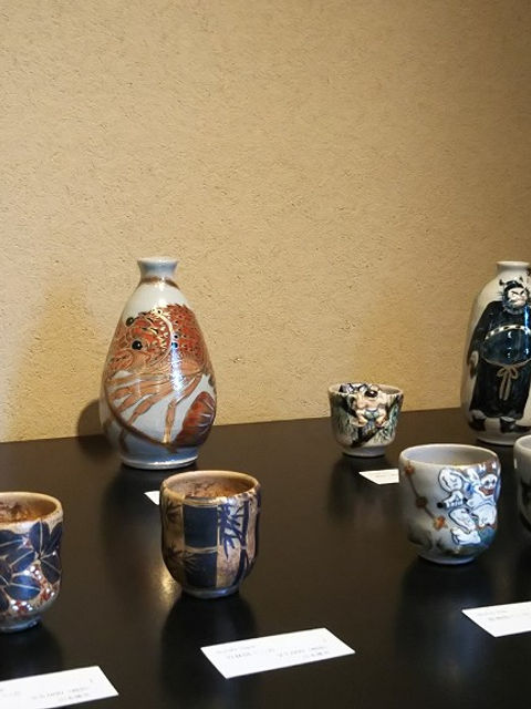 壁を背に黒い台に並べた酒器。徳利、ぐい呑、瓢箪形の瓶など