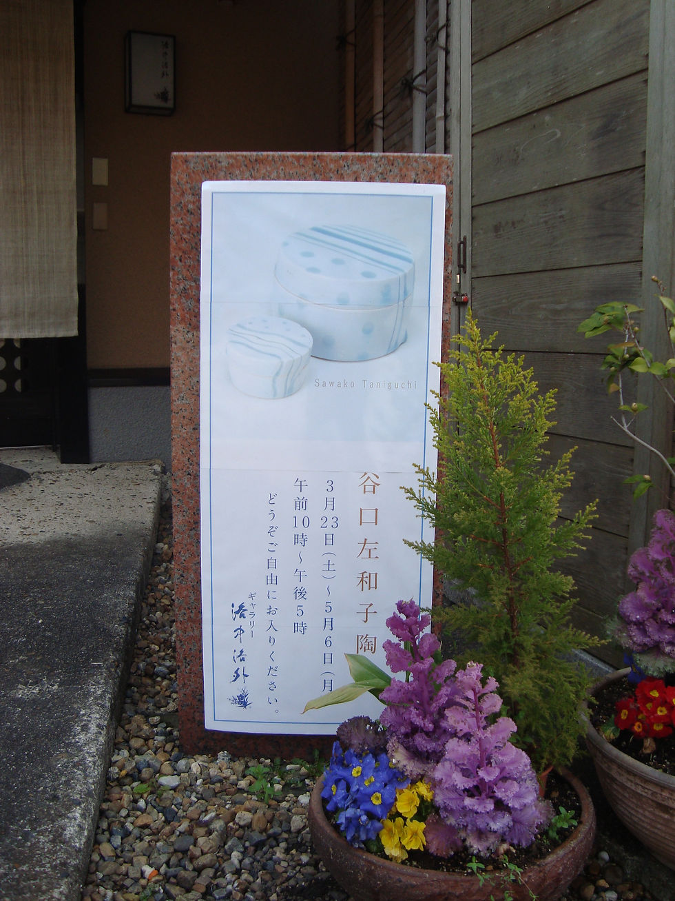 「谷口左和子陶展」の看板と、手前に咲くパンジーなど花の寄せ植え