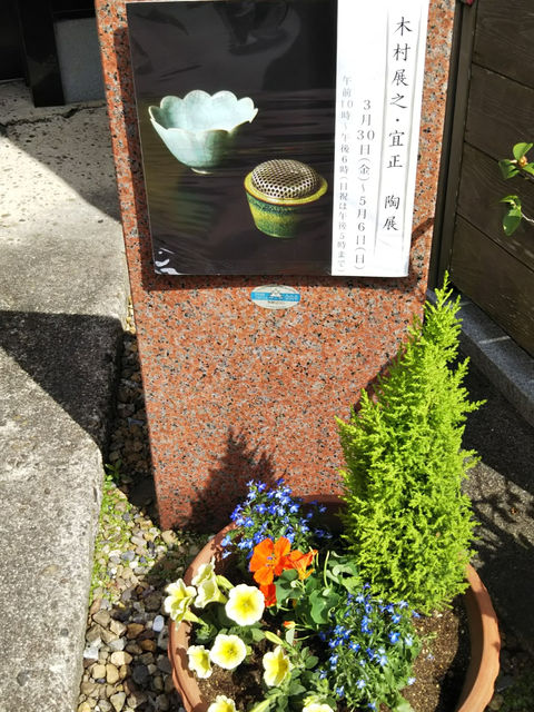 陶二人展の案内を掲げた石の看板と、足元に咲く黄色い花の寄せ植え
