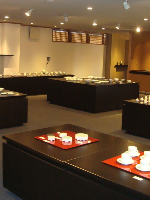 白磁の器が黒い台に並ぶ陶展の会場、赤い盆にカップをのせた手前の展示