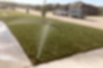 Residential lawn irrigation system watering an established cool season lawn in a suburban neighborhood during early morning hours. Pop-up sprinkler head applying deep, even coverage to promote healthy root growth, drought resistance, and improved lawn health in Southwest Missouri. Jasper Ridge landscaping trailer and work truck visible in the background.