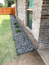 Decorative black rock foundation bed with small shrubs along a brick home.
