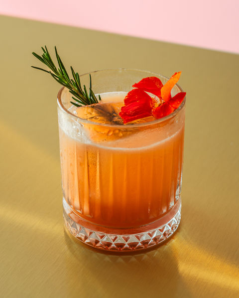 A deep orange cocktail, garnished with rosemary and a red flower, served on a gold bar.