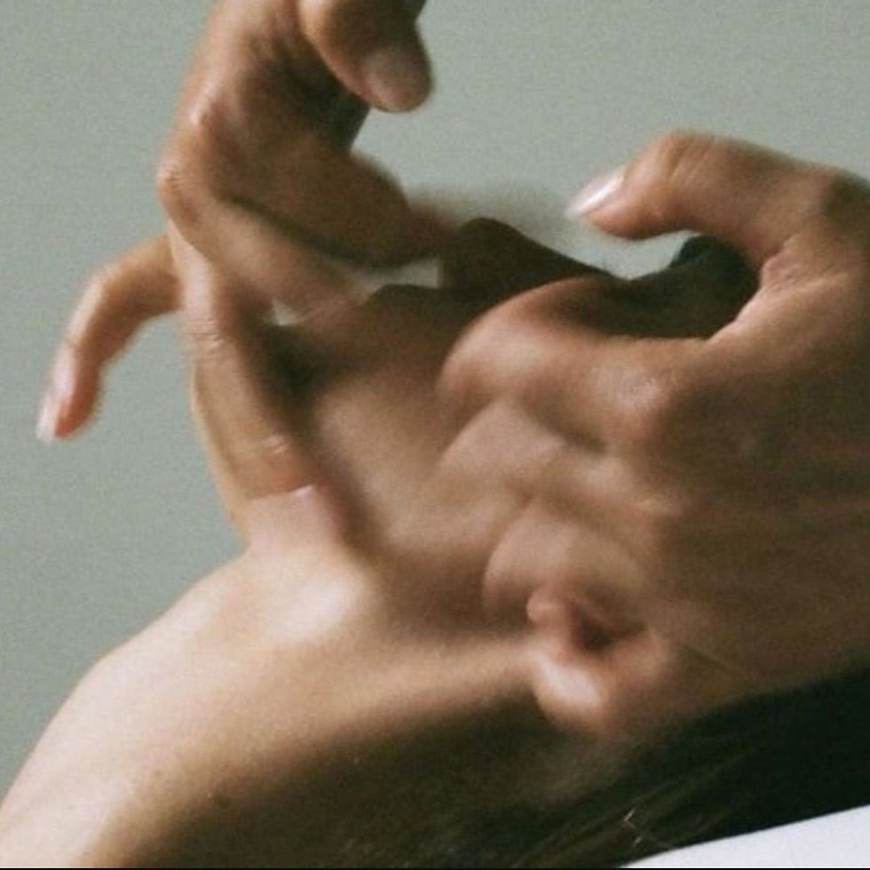 Close-up of two hands forming a heart shape over a bare chest against a plain gray background.