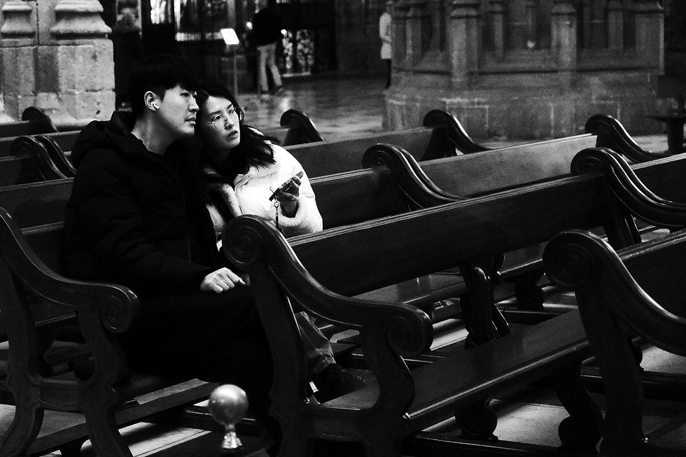 A couple sits closely on wooden pews in a dimly lit cathedral. They look attentive and contemplative, with intricate stone columns behind.