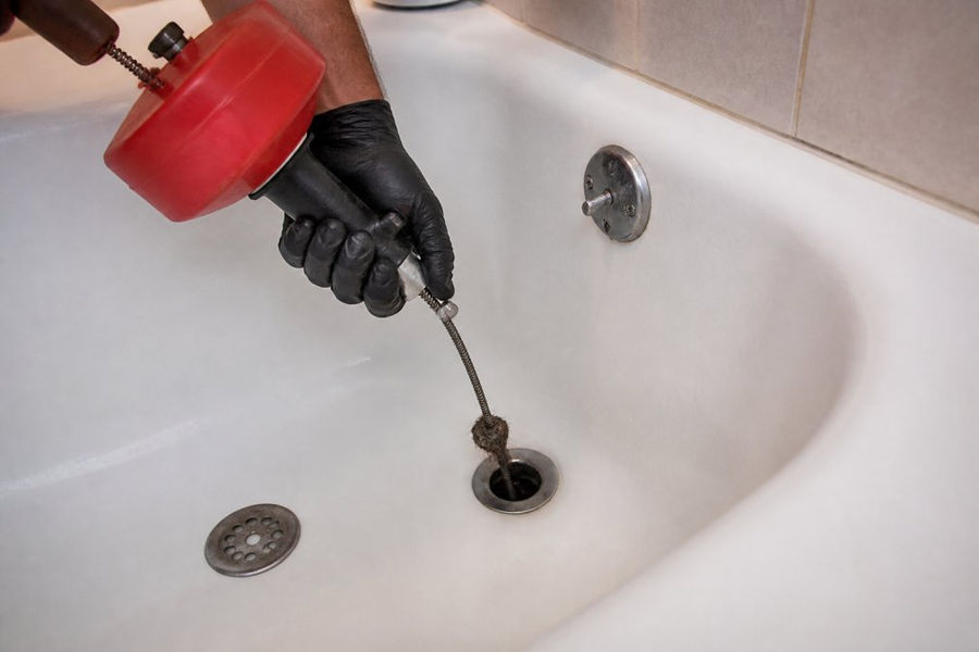 Close-up of a hand using a drain snake to remove debris and fix recurring drain clogs in a bathtub.