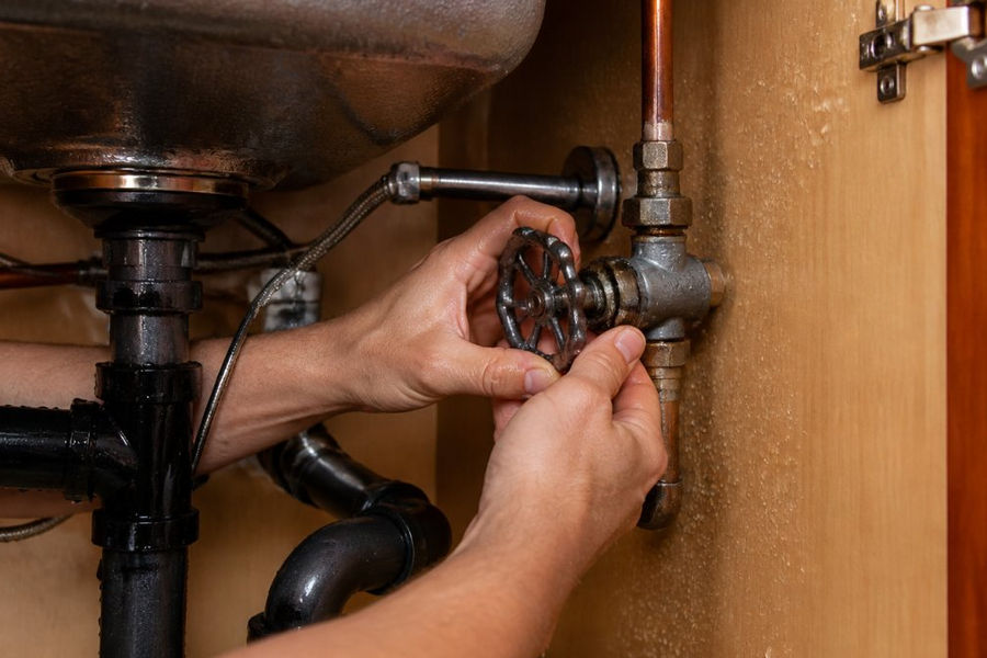 Homeowner turning off the main water valve under a sink to book emergency pipe repair online.