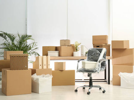 Packed office with moving boxes, desk, plants, and wrapped chair ready for relocation.
