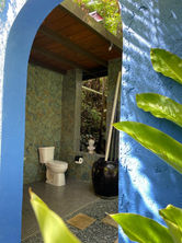 Entrance of the private tropical bathroom of the Bali Cabin at Mont Carpe Diem, featuring an open-air shower surrounded by lush greenery and covered toilet and sink. 