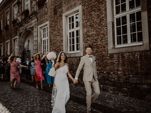 glückliches Brautpaar läuft Hand in Hand und hinter ihnen laufen ihre Gäste. Braut trägt Schleier und modernes enges Brautkleid