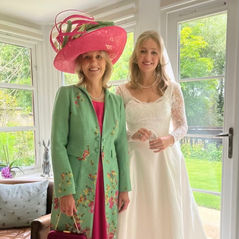 Mother of the Bride and her daughter just about to leave for the wedding wearing a grass-green linen long coat which has embroidered flowers and leaves 'growing' up from the hem, over a plain raspberry pink dress and very large 'picture hat' with an upturned brim and trimmed with lots of curled dyed ostrich feathers peeping up from the behind the brim.