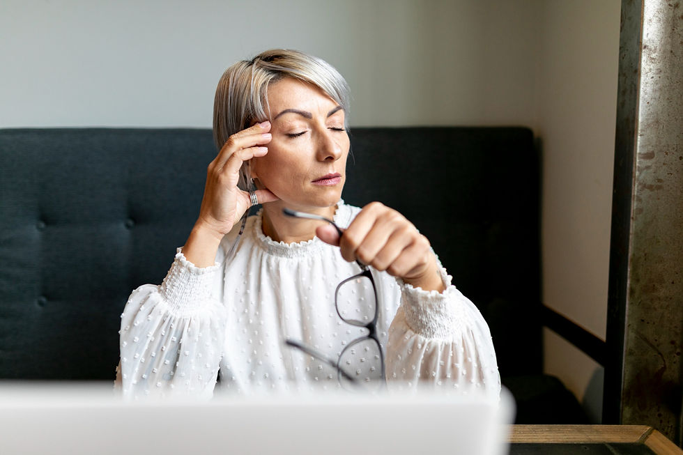 Professional woman with short gray hair holding glasses and touching her temple, appearing fatigued or mentally drained while working on a laptop.