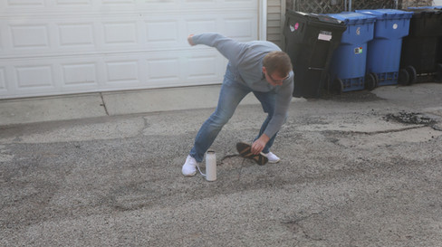 A man in an alley grabbing a gym shoe near a metal water bottle mid-run. The background includes garbage cans and a garage door