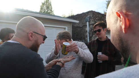 A man taps out the last drops of a Twisted Tea into a bottle cap held by another man as others observe